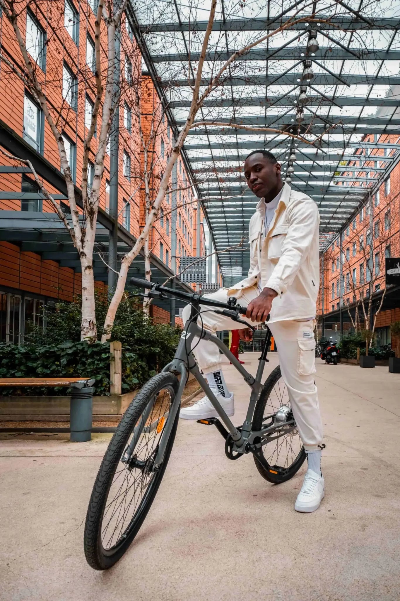 Stylish man in white outfit posing with black Ref Essential bike against a modern building backdrop, showcasing urban lifestyle.