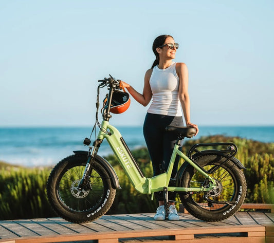 Woman standing with Eskute Star folding e-bike on a boardwalk overlooking the ocean, highlighting the portability and style of green urban electric bicycles for eco-friendly commuting.