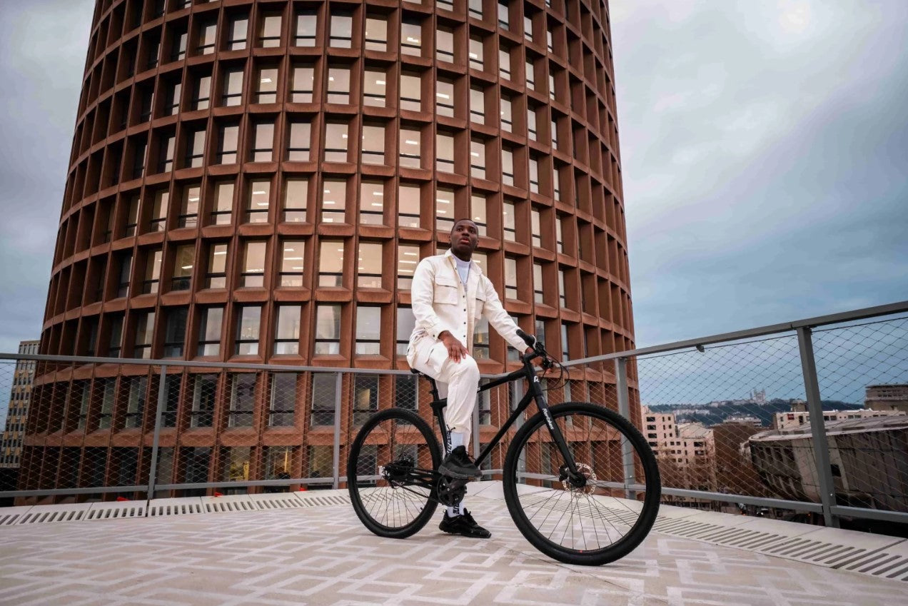 Stylish man in white outfit posing with black Ref Essential bike against a modern building backdrop, showcasing urban lifestyle.