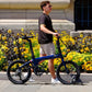 Active man walking with a folded blue Eole X ebike on a sunny city sidewalk lined with vibrant yellow flowers.
