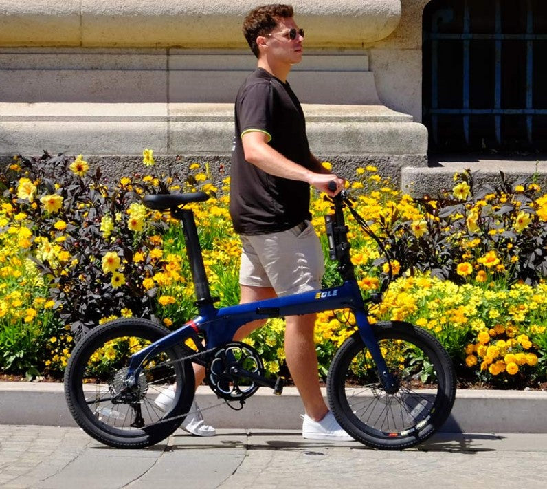 Active man walking with a folded blue Eole X ebike on a sunny city sidewalk lined with vibrant yellow flowers.