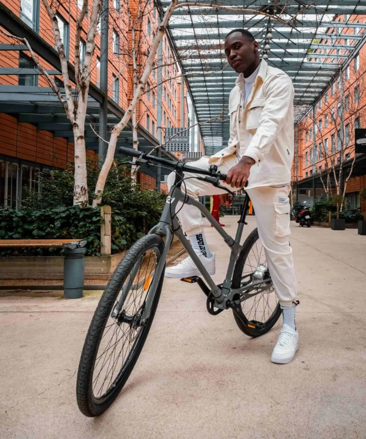 Young man standing with a dark Essential Boost bike, in a modern urban courtyard.