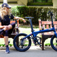 Relaxed woman seated next to a parked blue Eole X ebike, representing the leisurely and stylish aspect of electric cycling in urban parks.