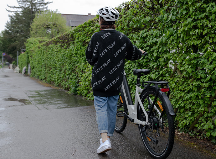 Person walking with a Deruiz Quartz e-bike on a leafy sidewalk, showcasing real-life use.