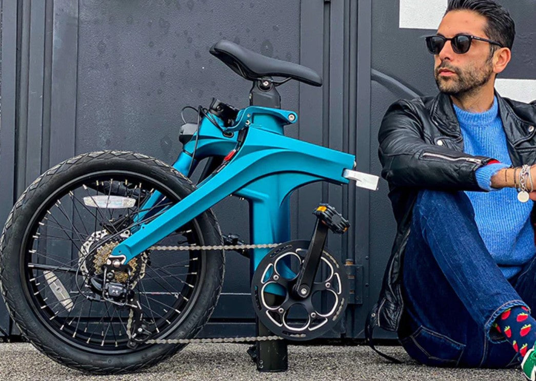 Man sitting next to the blue Fiido X electric bicycle showing its foldable features and compactness.